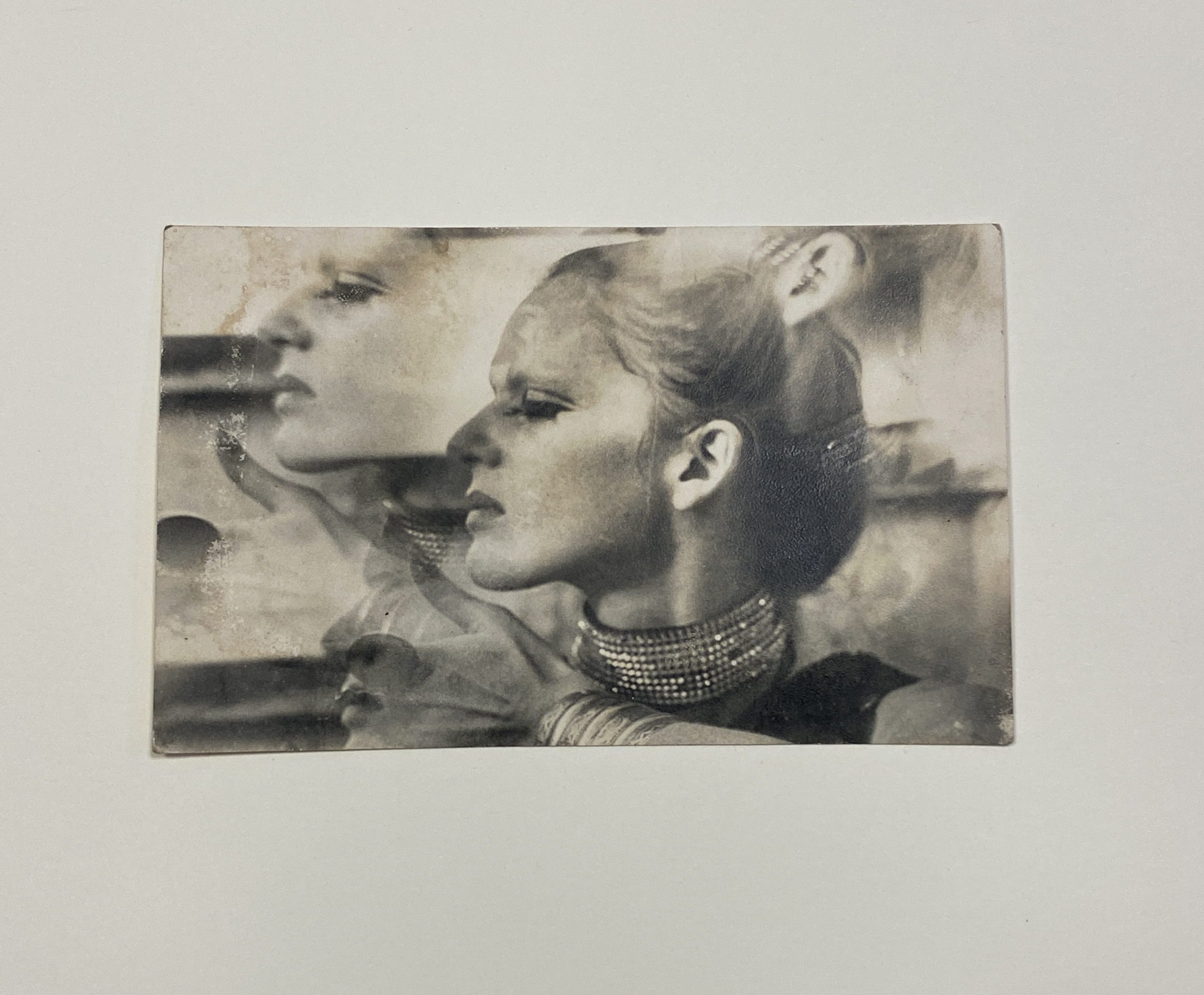 Black and white double-exposed photograph with double exposure of a blonde woman's face in profile. She is wearing a jewelled choker and bracelets, and holding up her left thumb.