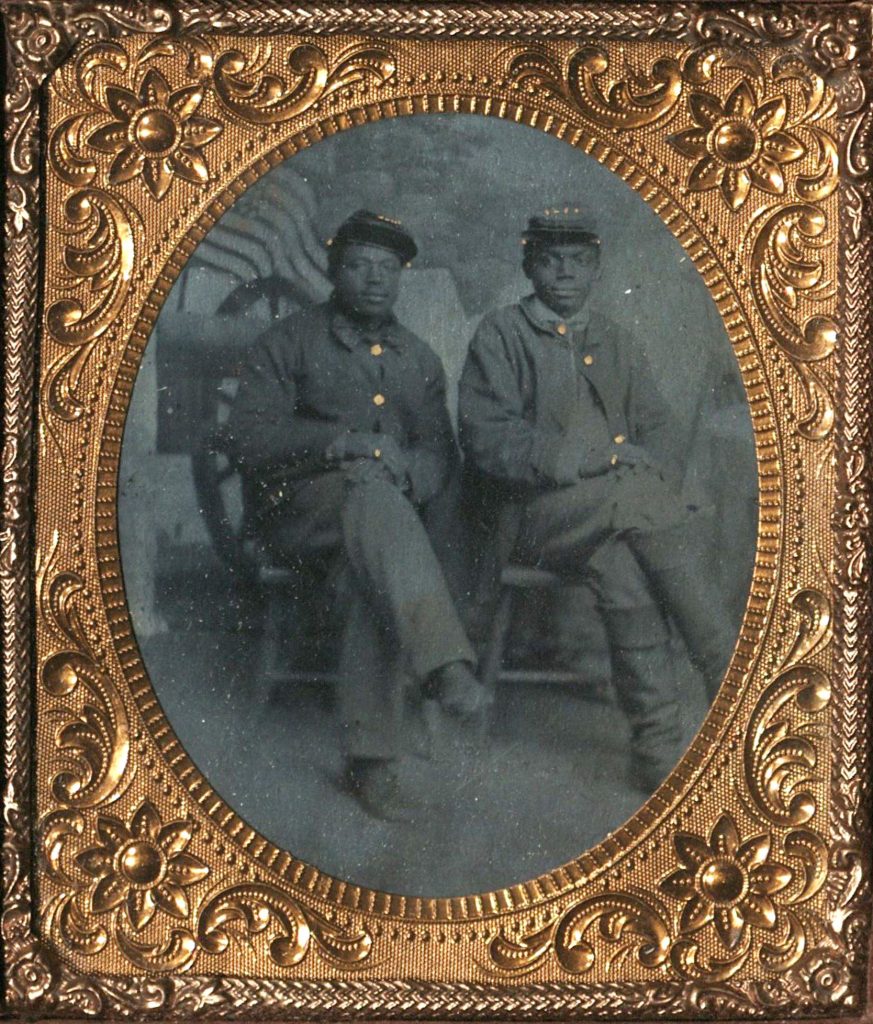 Black and white tintype portrait of two Black soldiers in uniform seated; American flag in background. Tintype portrait is encased in ornate gold frame. 