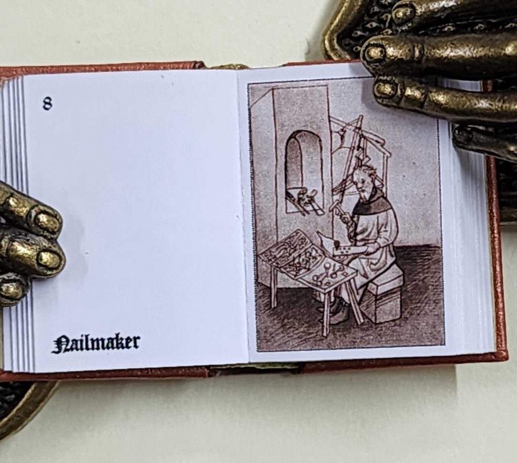 Miniature book is held open to show a Medieval Nail maker at work.