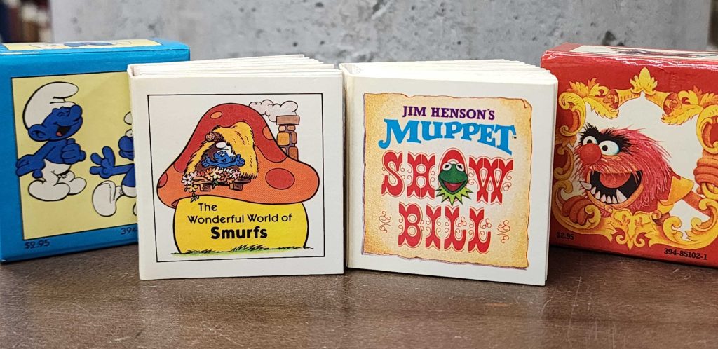 Two miniature books pictured. On the left: The wonderful world of Smurfs with a blue slipcase with two smurfs laughing. On the right: Jim Henson's mupper show bill with a red slipcase depicting Animal.