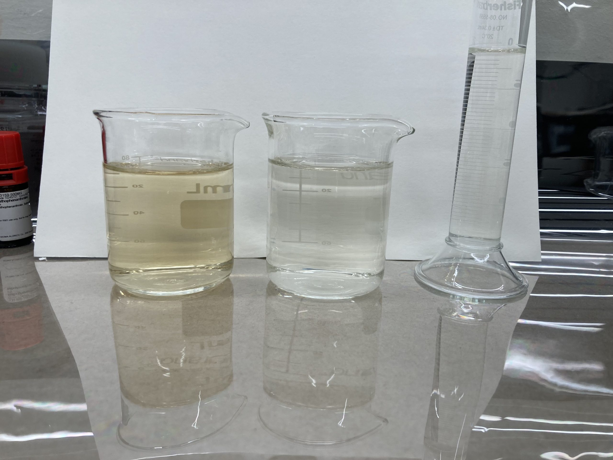 Three beakers containing water are shown against a white paper background. The first beaker is quite yellow, the second beaker is slightly yellow, and the third beaker appears almost completely clear.