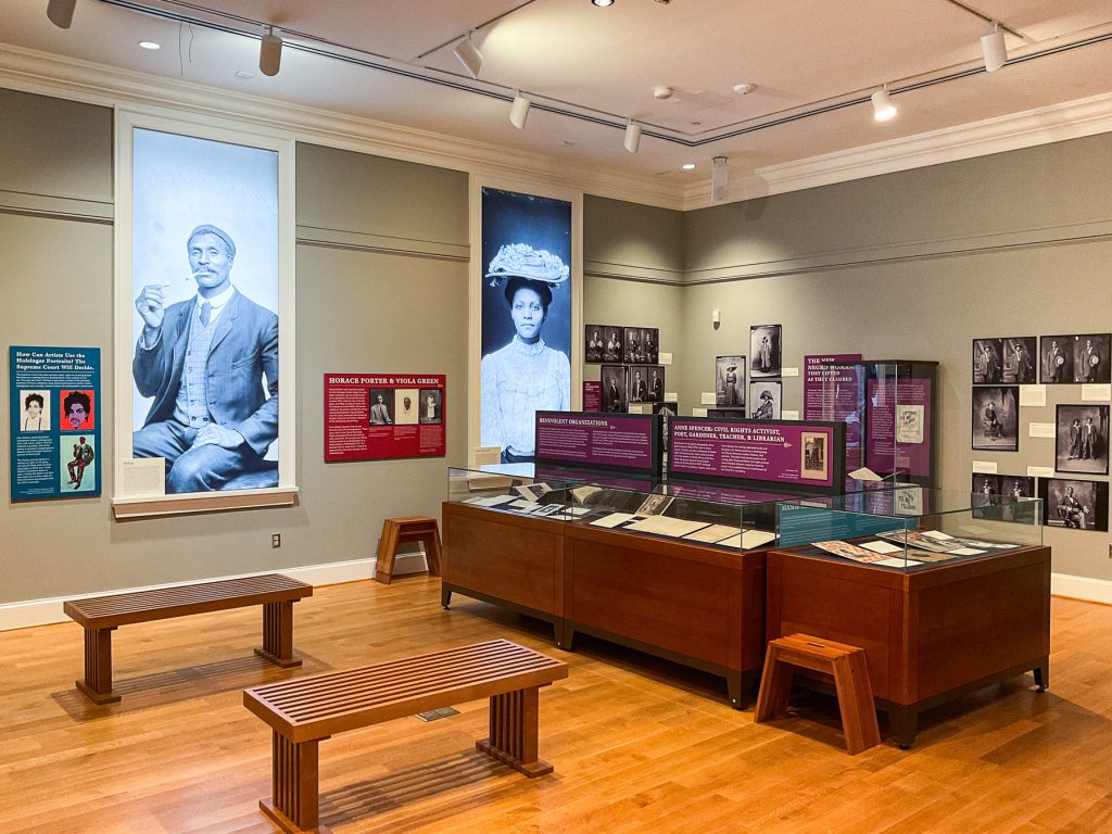 Gallery view showing wall-mounted photographs, backlit photographs in windows, and display cases featuring African American portraits from the Holsinger Studio Collection. 