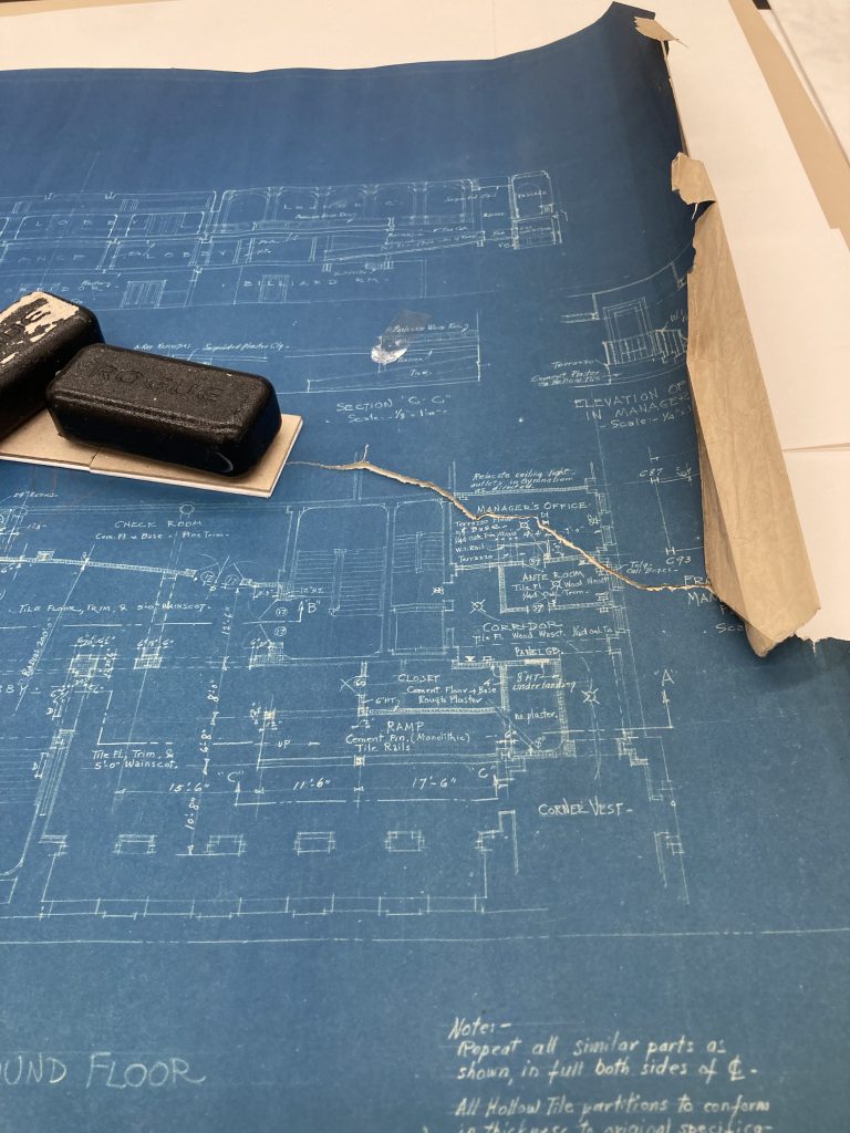 A blueprint is being mended, with a stack of board weight down with a black weight on top of a torn section of the blueprint.