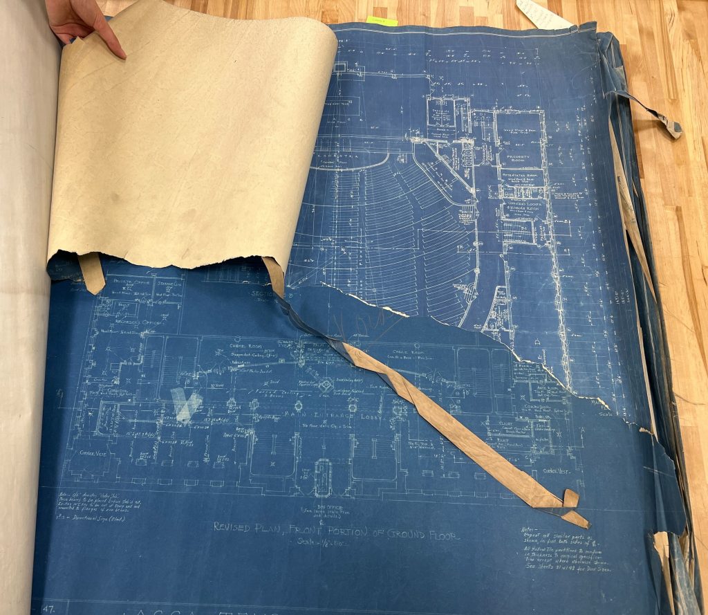 Image of a blueprint showing the inside of a building. A hand at the top left of the image is holding back half of the blueprint to demonstrate it has been torn in half. 