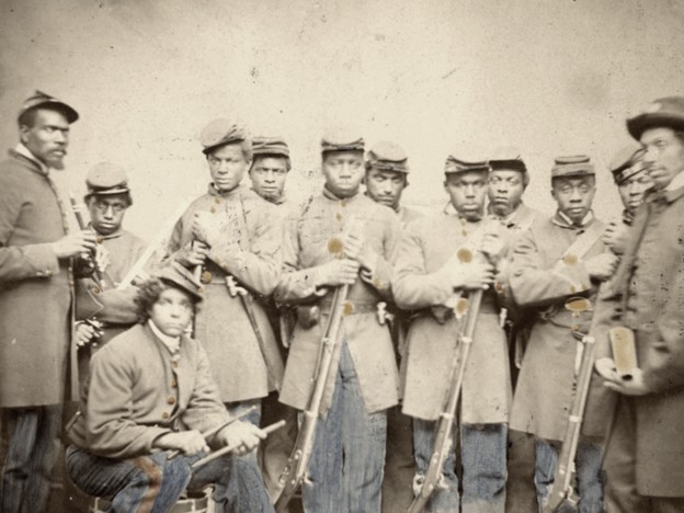 Sepia-toned Civil War era photograph of approximately ten African American Union soldiers in Union Army overcoats and kepi caps, most holding rifles upright. One soldier crouches in the front center. The men face the camera with serious expressions. Private Samuel Bond of Company C, 19th USCT, is pictured center sixth from the left.
