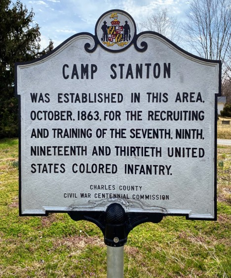 Historic marker, Black text on white background on a post on the site of Camp Stanton with the Charles County Civil War Centennial Commission seal at the top of the sign.