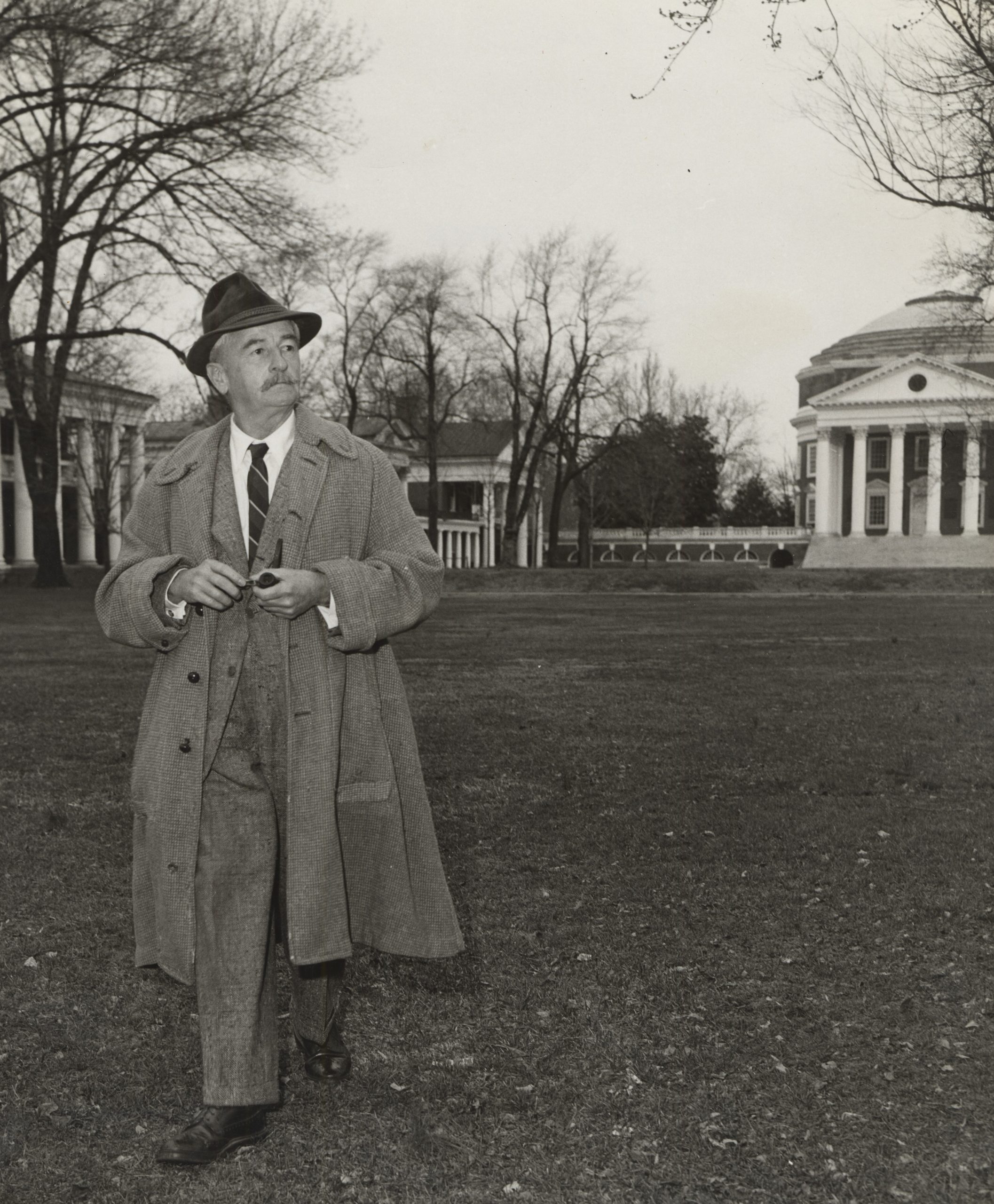 Faulkner looking off to the side while walking in front of the rotunda on UVA Grounds wearing a suit, tie, hat, and jacket and holding pipe