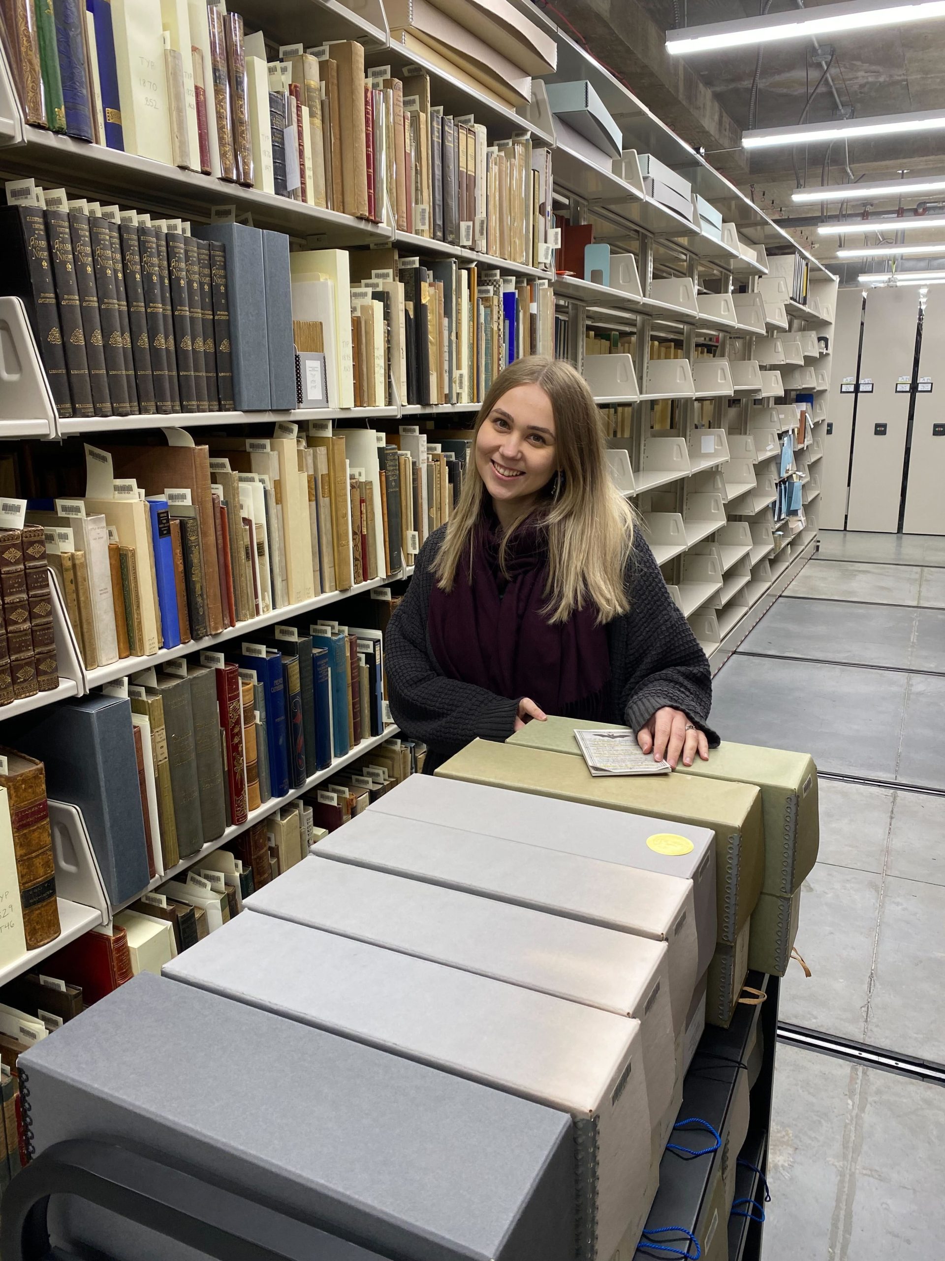 smiling blond woman surrounded by shelves of books and posed with a book truck of archival boxes