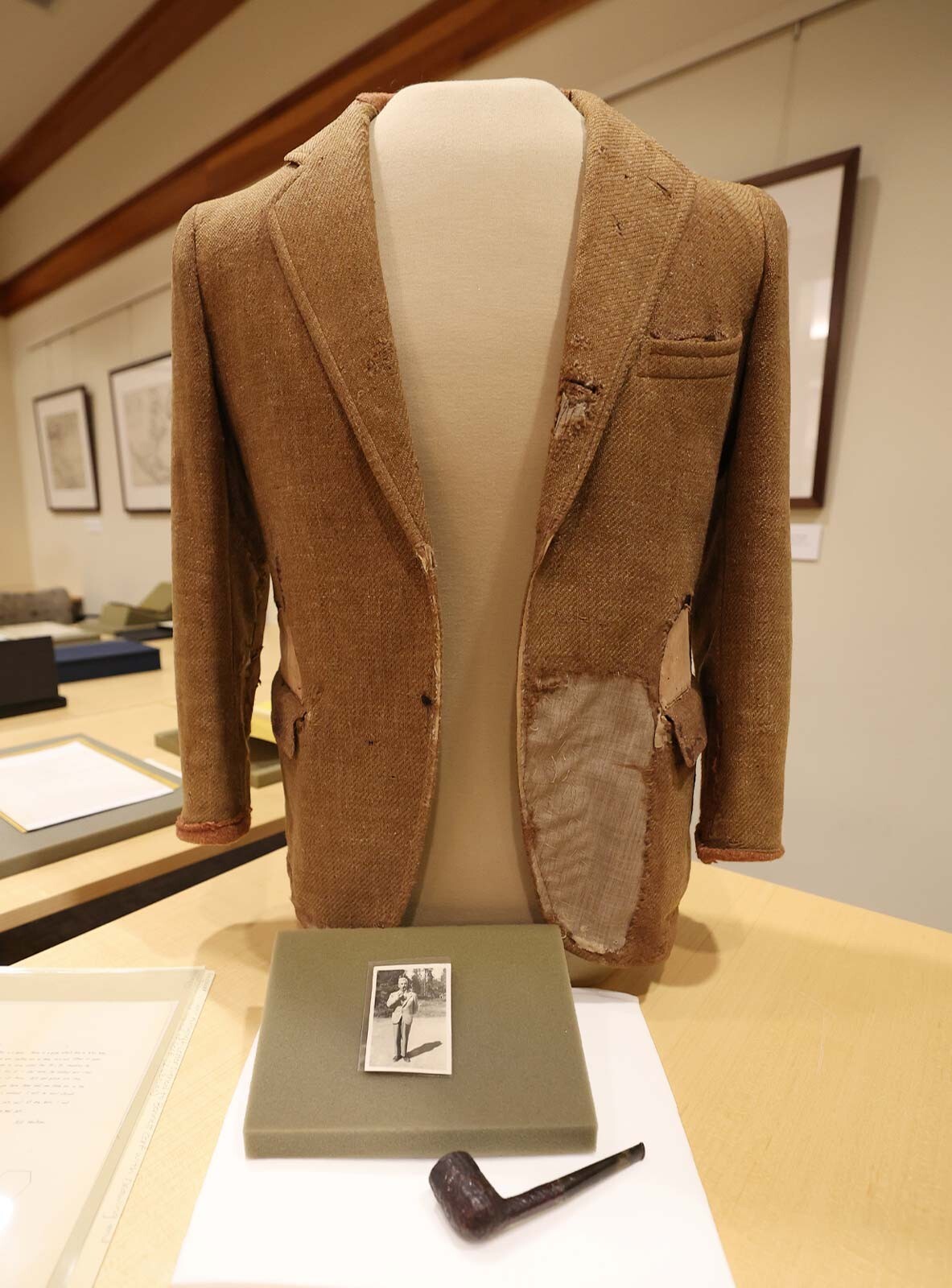 a well-worn beige tweed jacket propped onto a mannequin torso and posed behind a brown pipe alongside a photograph of Faulkner wearing the jacket and smoking the pipe